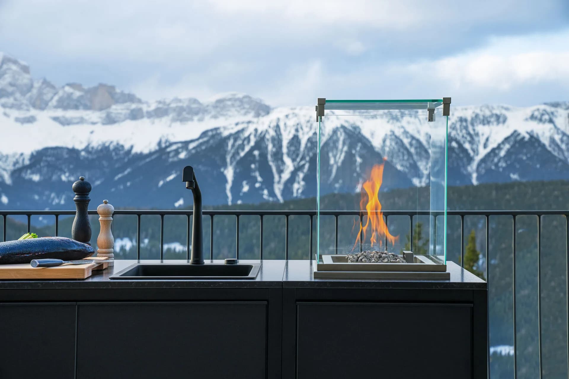 Detail einer dunklen Outdoor-Küche mit schwarzer Spüle und offener Feuerstelle hinter Glas, im Hintergrund verschneite Berge