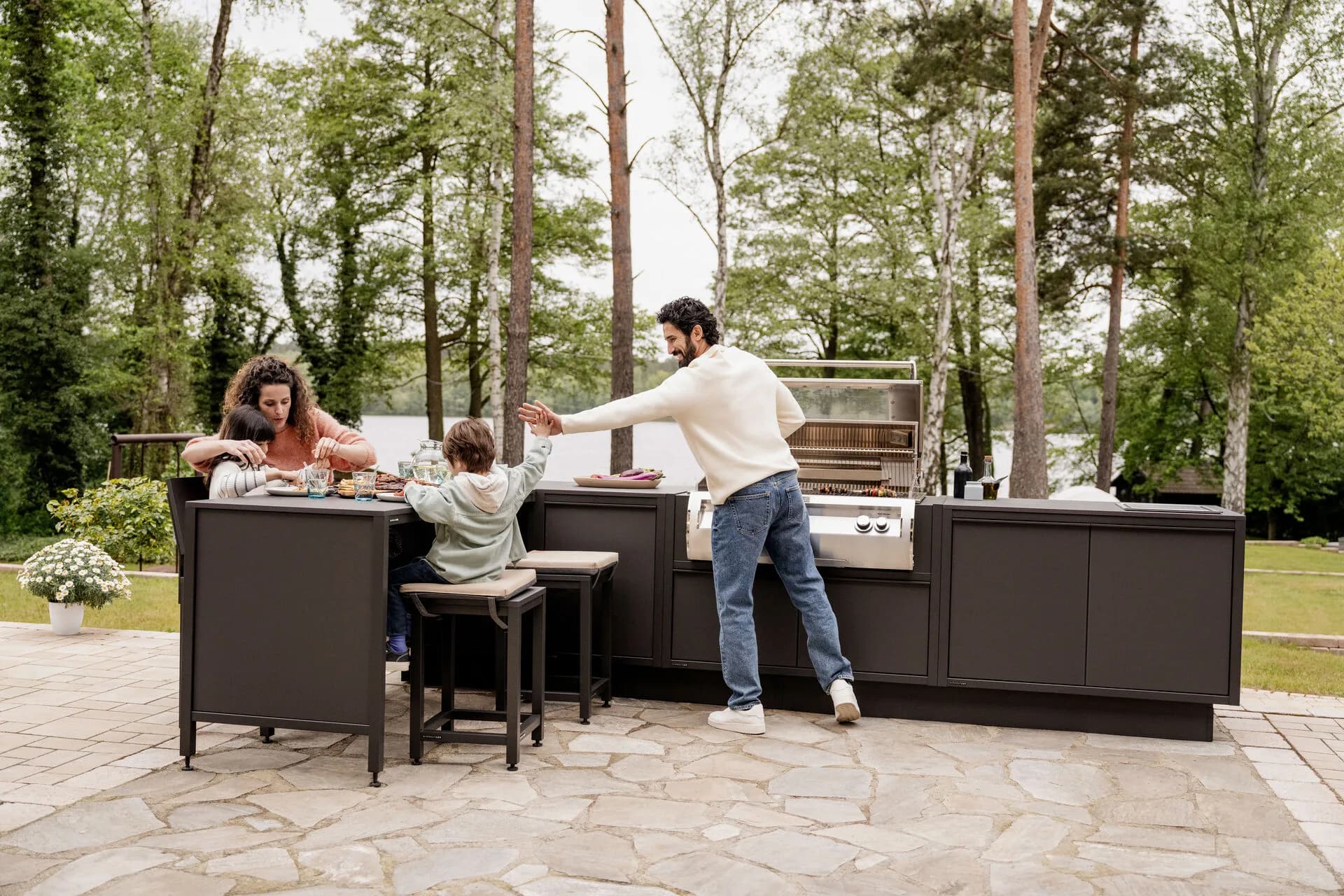 Familie sitzt und grillt an einer anthrazitfarbenen Outdoor-Küche mit Sitztheke auf einer Natursteinterrasse am Waldrand