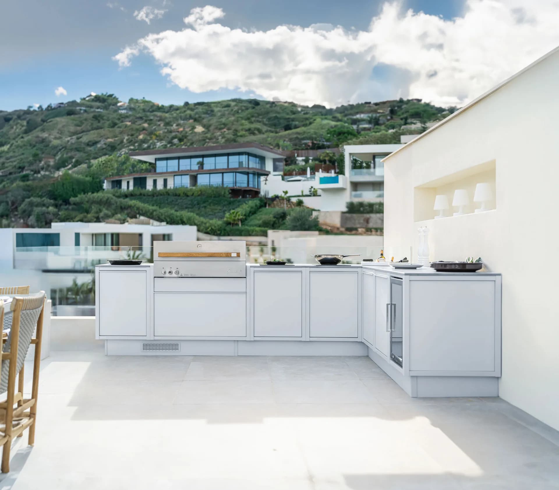 Helle Outdoor-Küche mit Edelstahlgrill auf einer sonnigen Terrasse, dahinter ein Hang mit Villen und das Meer