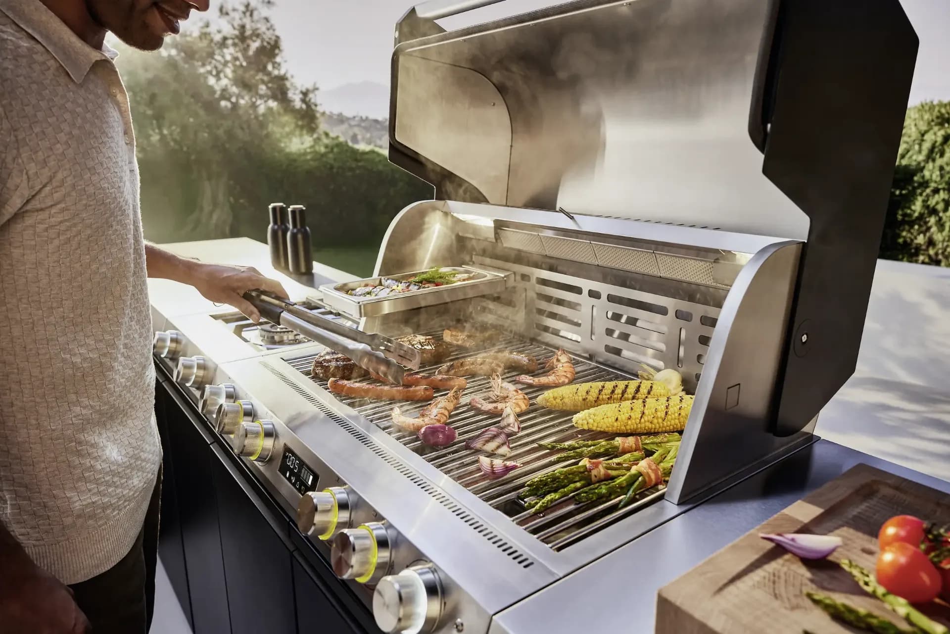 Mann grillt Steaks, Garnelen, Mais und Gemüse auf einem großen Miele-Edelstahlgrill mit geöffneter Haube, im Hintergrund ein Garten