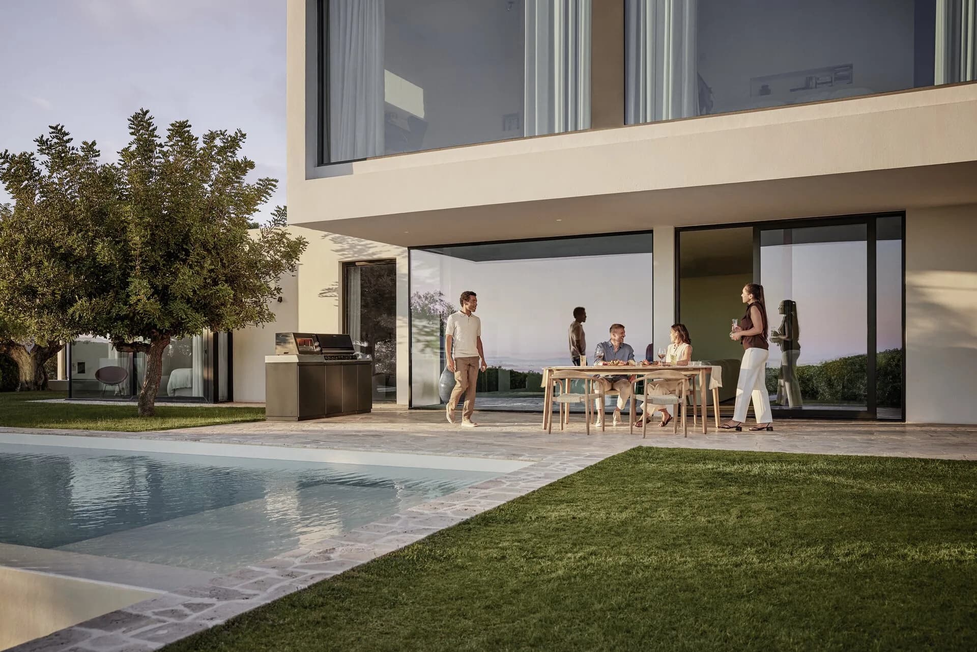 Freunde essen in der Abenddämmerung an einem Holztisch auf der Terrasse einer Villa, daneben eine Miele-Outdoor-Küche am Pool