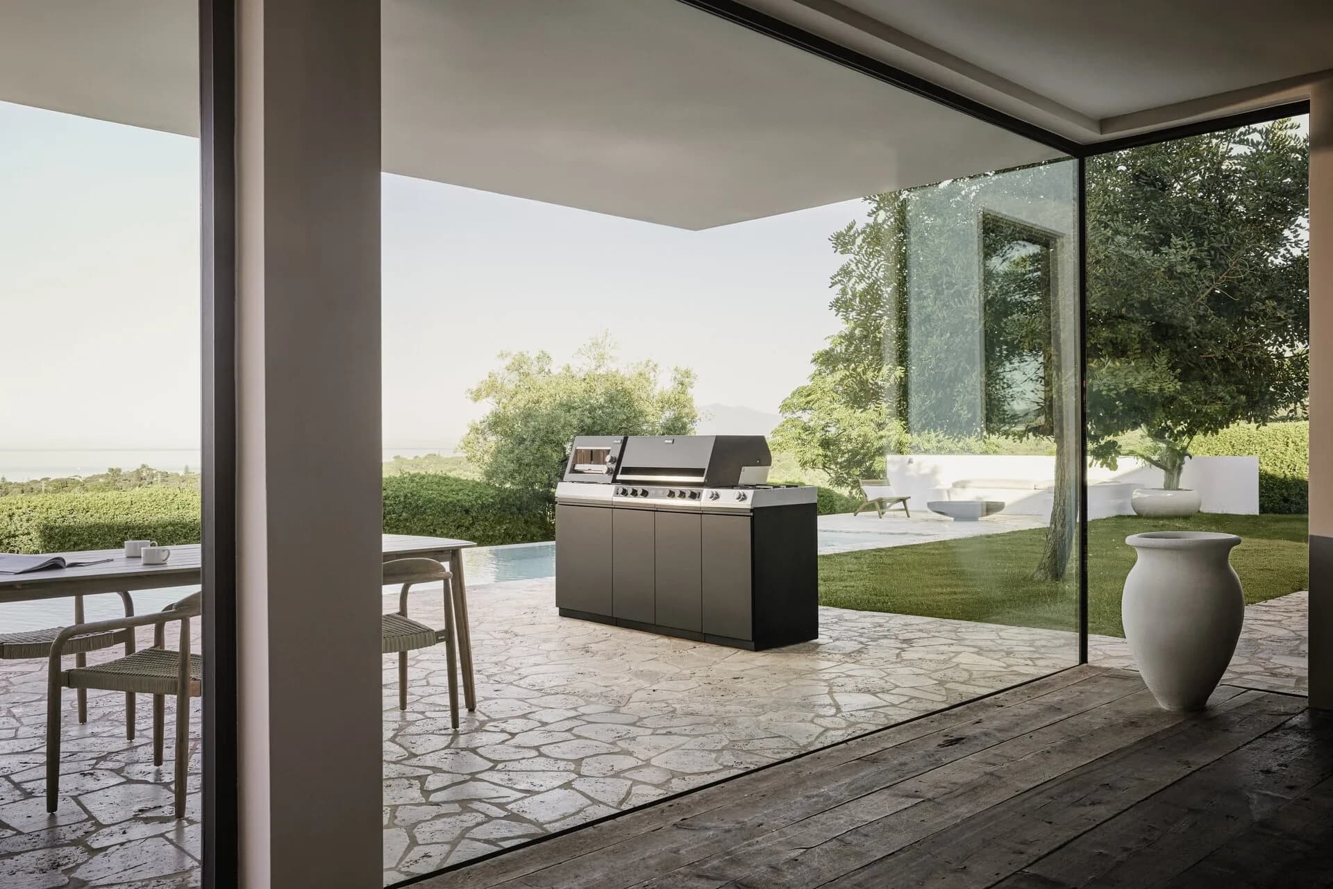 Freistehende Miele-Outdoor-Küche auf einer Natursteinterrasse mit Blick auf Pool, Garten und Meer, gesehen durch eine große Glasfront