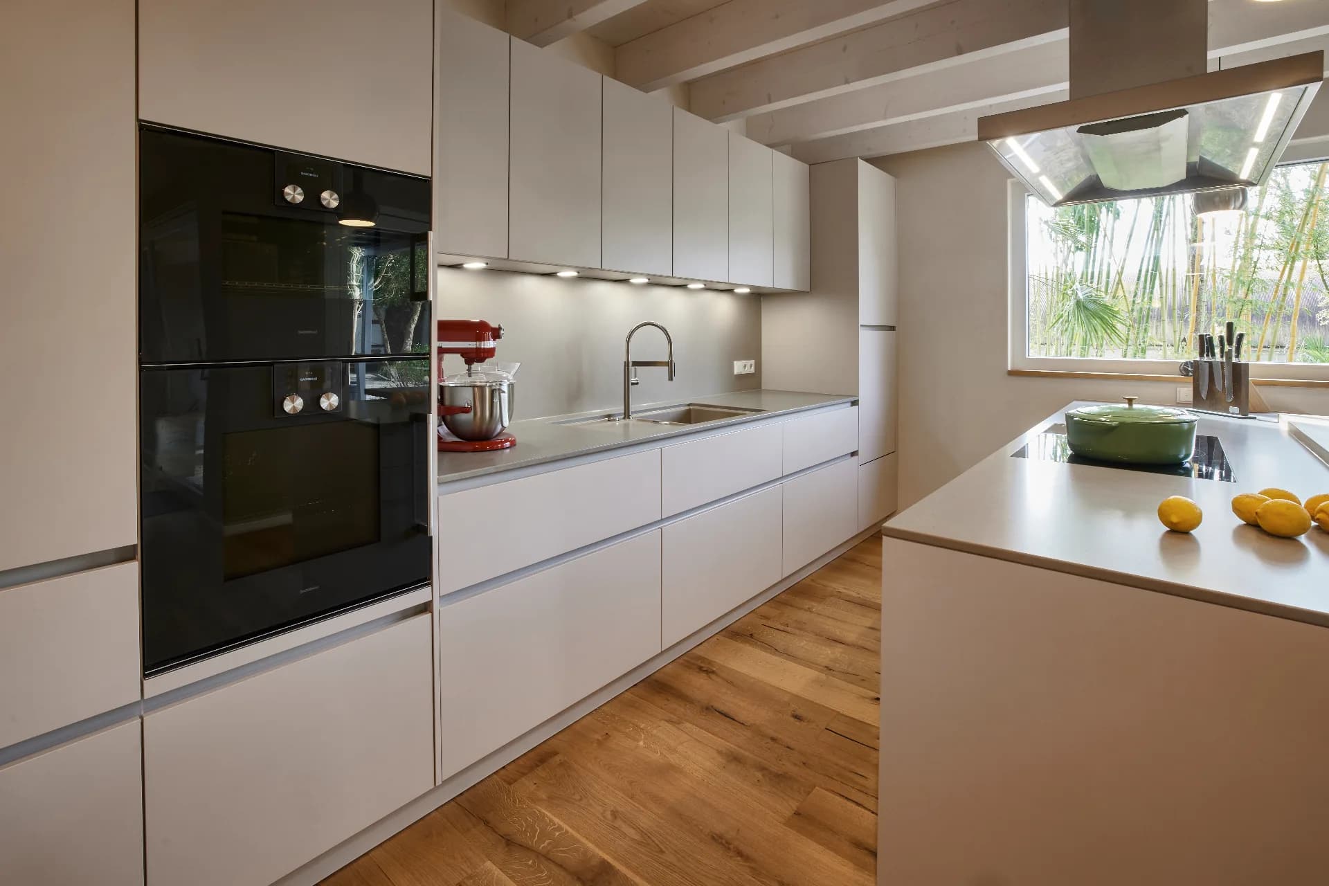 Küche mit greigen Fronten, Gaggenau-Backöfen und Kochinsel unter einer Edelstahlhaube