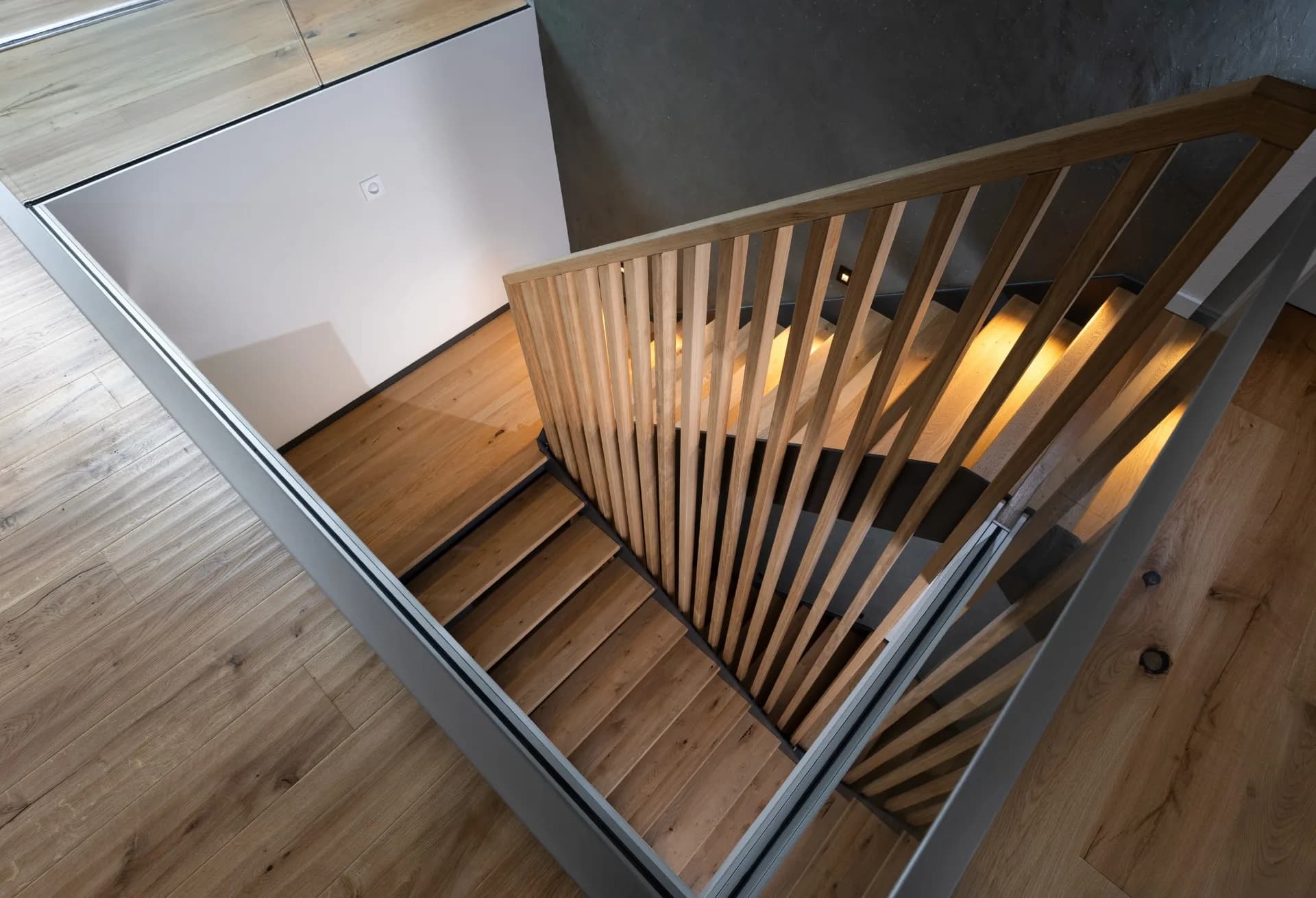 Blick von oben auf eine halbgewendelte Treppe mit Geländer aus Eichenleisten und beleuchteten Eichenstufen vor dunkler Wand