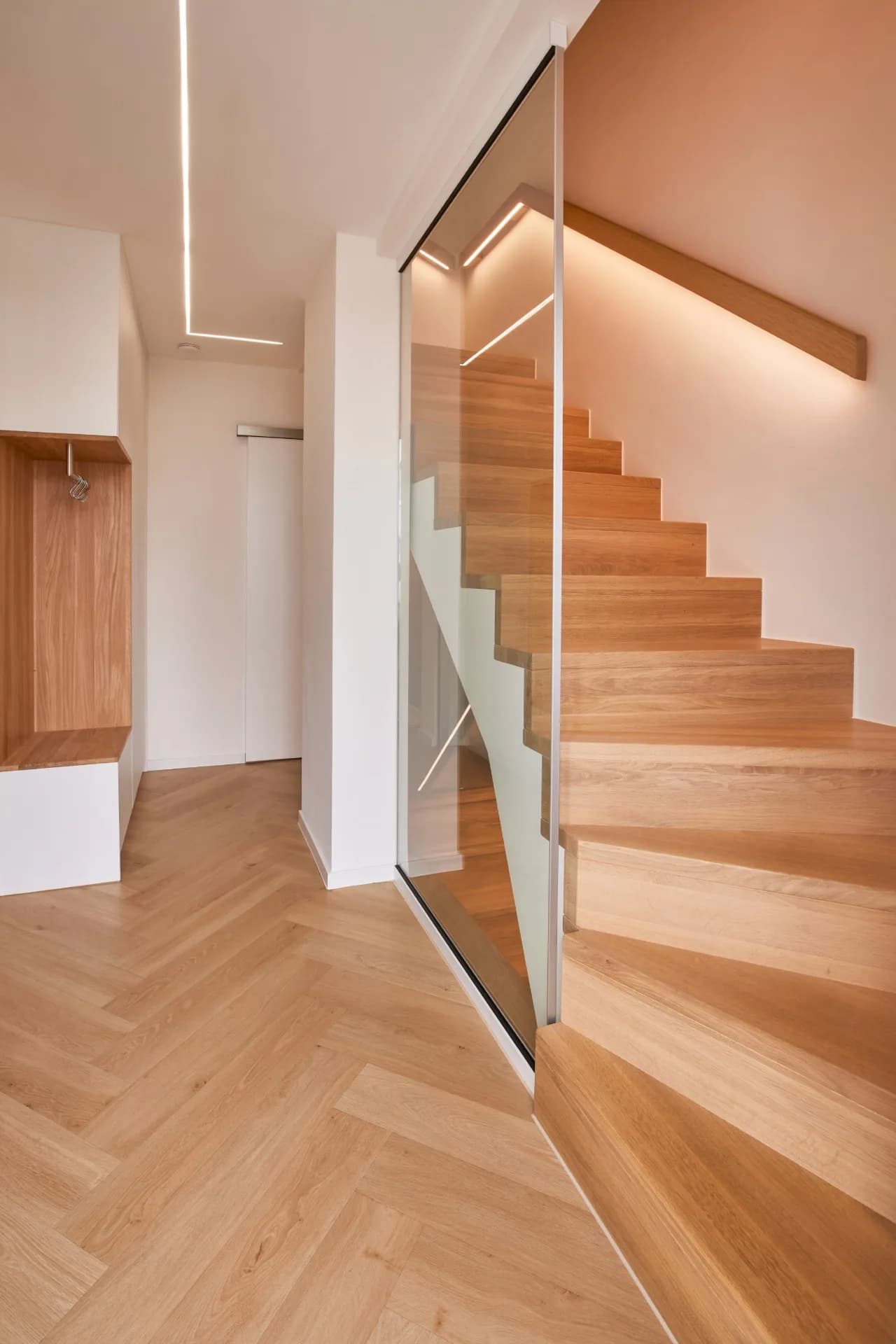 Gewendelte Betontreppe, belegt mit Stufen aus Eiche, hinter einer Glaswand in einem Flur mit Fischgrätparkett